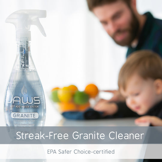A bottle of Jaws Granite Streak-Free Granite Cleaner is shown in the foreground on a white background, with a blurred lifestyle scene in the background of a man and a child looking at his reflection in counter