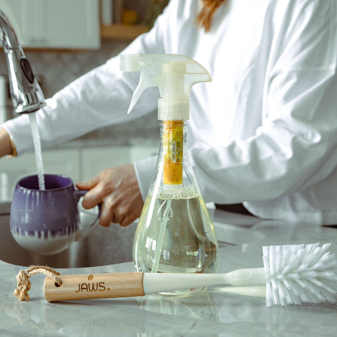 JAWS Dish Spray and brush on counter with woman rinsing a mug in the background.