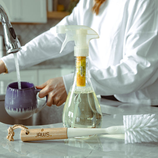 JAWS Dish Spray and brush on counter with woman rinsing a mug in the background.