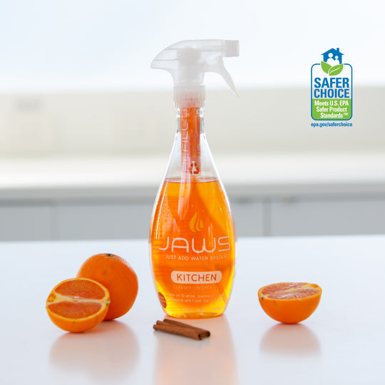Bottle of JAWS Kitchen Cleaner on reflective surface with orange section and cinnamon next to it.  EPA Safer choice logo in background.