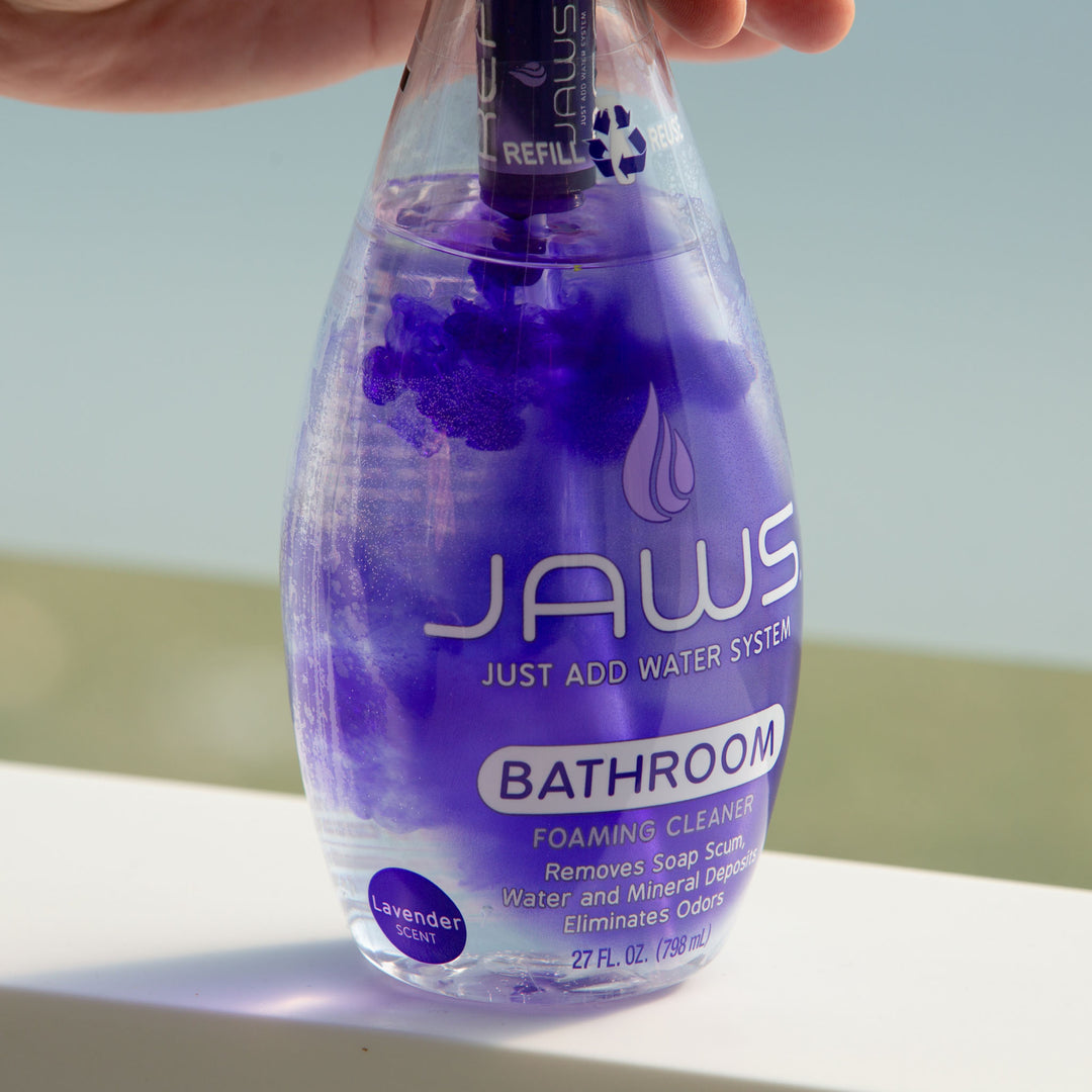 A hand holding a bottle of JAWS Foaming Bathroom Cleaner, with purple JAWS liquid, set against a blurred background.