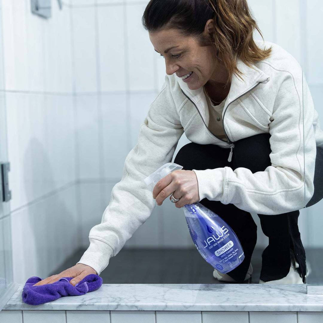 Woman cleaning a bathroom shower with a purple microfiber cloth and a bottle of JAWS Shower Cleaner.