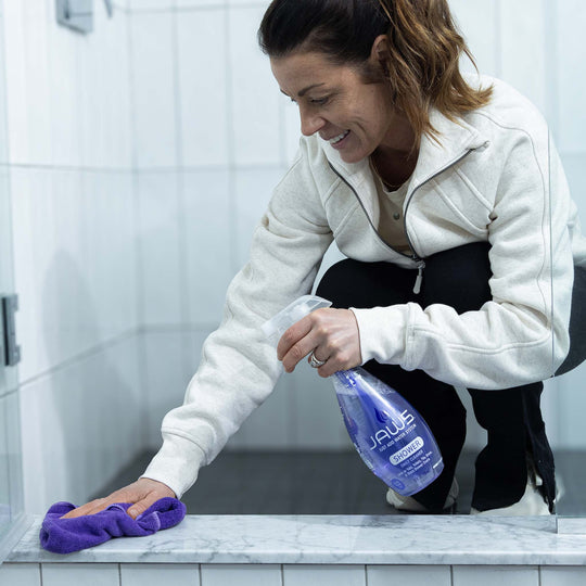 Woman cleaning a bathroom shower with a purple microfiber cloth and a bottle of JAWS Shower Cleaner.