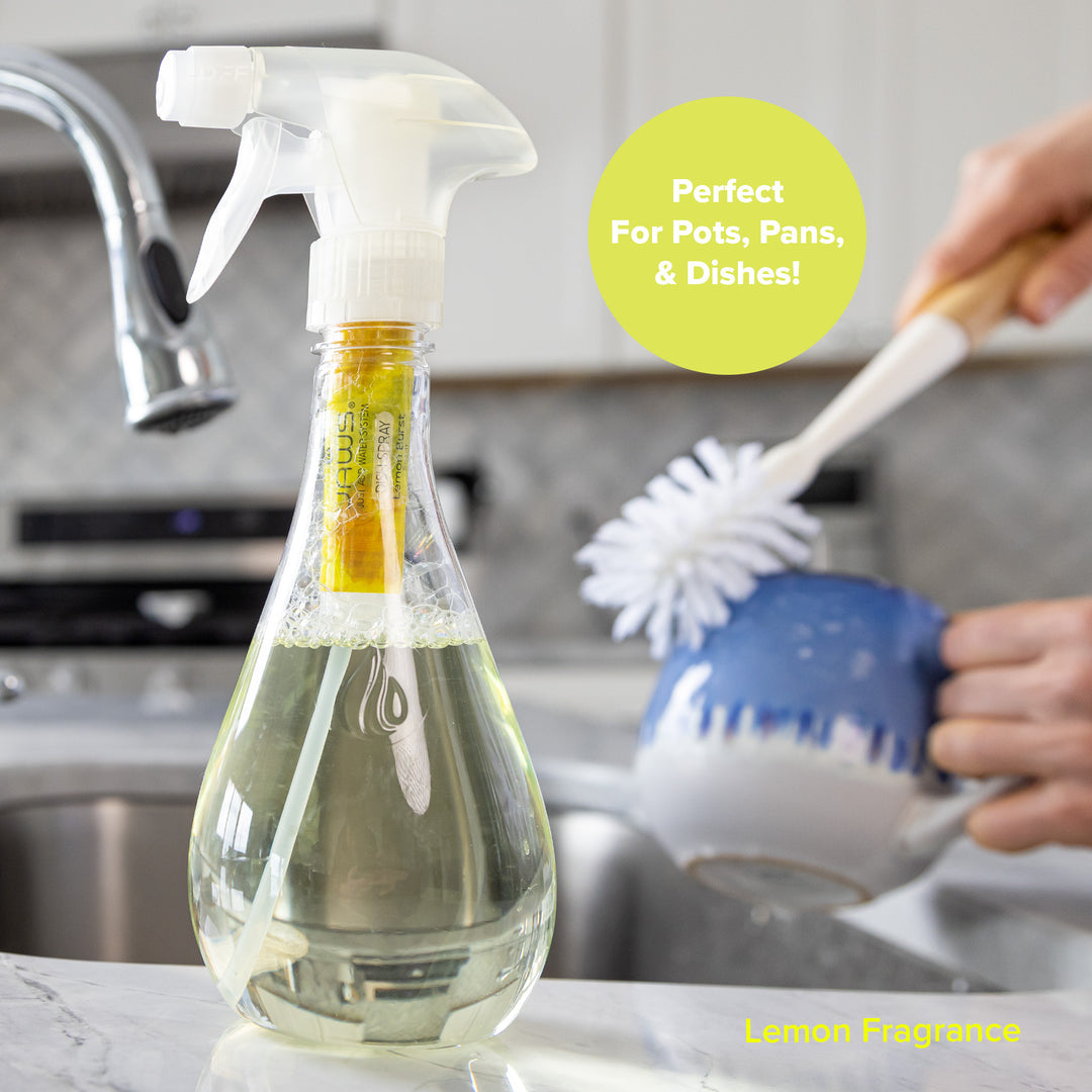 A clear bottle of JAWS Dish spray filled with light yellow dish soap, a white trigger sprayer and JAWS Refill in the neck of the bottle, shown in a kitchen setting with a hand using a JAWS dish brush on a blue-and-white mug. A yellow circle reads 'Perfect for Pots, Pans, & Dishes!' and the bottle label says, 'Lemon Fragrance'.