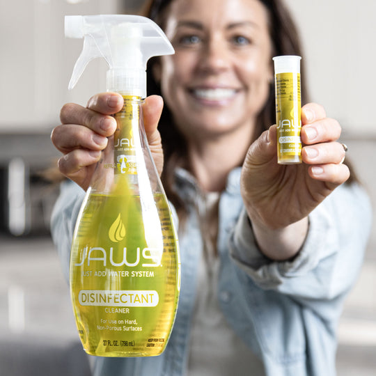 Woman holding a yellow disinfectant spray bottle and a Disinfect refill pod in a kitchen setting.