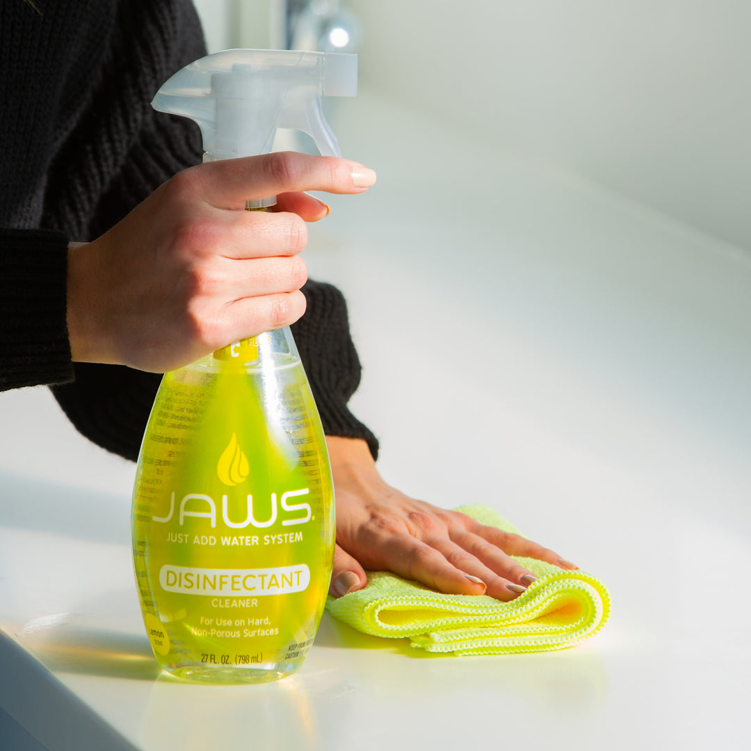 Person holding a JAWS disinfectant spray bottle with a yellow cloth on a light surface.