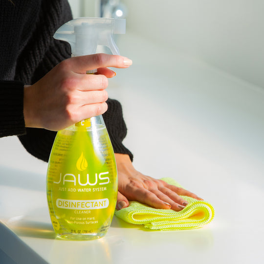 Person holding a JAWS disinfectant spray bottle with a yellow cloth on a light surface.