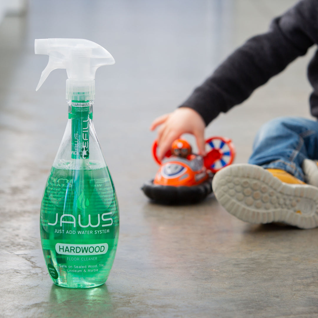 Bottle of JAWS hardwood floor cleaner on a polished wooden floor with a child playing in the background.