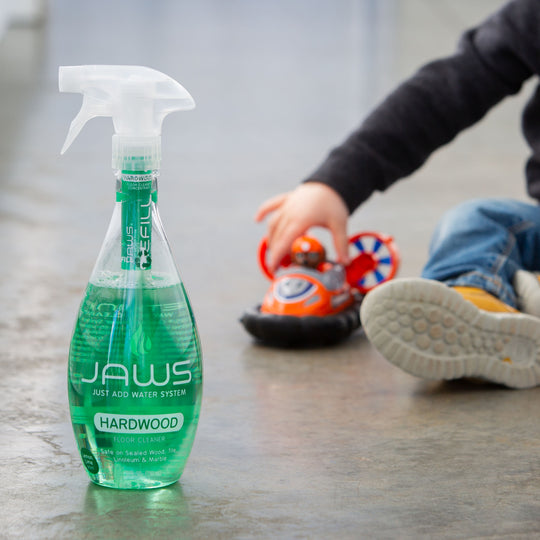 Bottle of JAWS hardwood floor cleaner on a polished wooden floor with a child playing in the background.