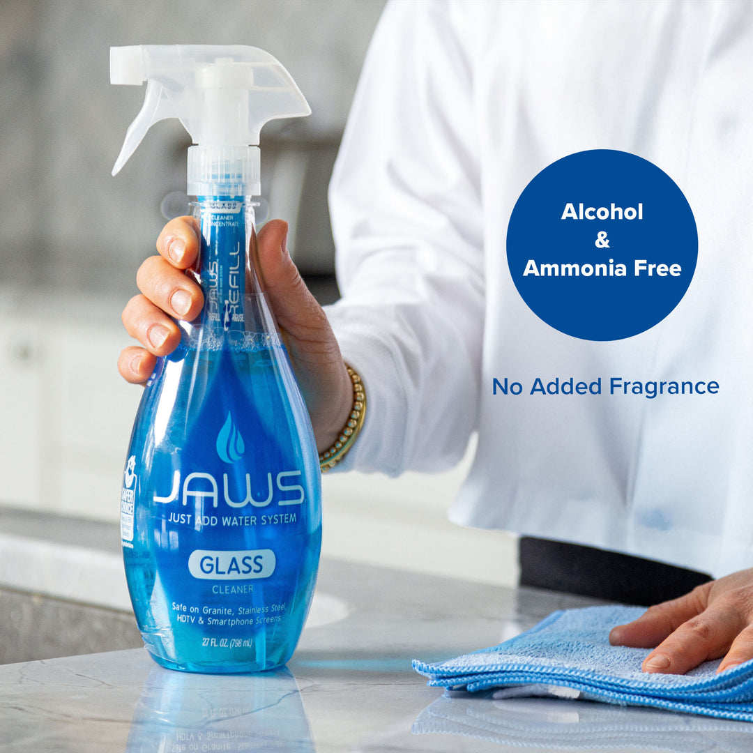 A person in a white shirt holds a blue trigger spray bottle of JAWS Glass Cleaner on a light countertop, with a blue microfiber cloth nearby and text overlays reading 'Alcohol & Ammonia Free' and 'No Added Fragrance'.