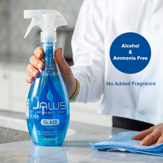 A person in a white shirt holds a blue trigger spray bottle of JAWS Glass Cleaner on a light countertop, with a blue microfiber cloth nearby and text overlays reading 'Alcohol & Ammonia Free' and 'No Added Fragrance'.