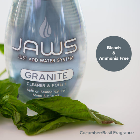 A bottle of JAWS Granite Cleaner & Polish from the Just Add Water System, next to fresh green basil leaves on a white surface. A dark gray circular badge on the right reads 'Bleach & Ammonia Free' and 'Cucumber/Basil Fragrance' is printed at the bottom.