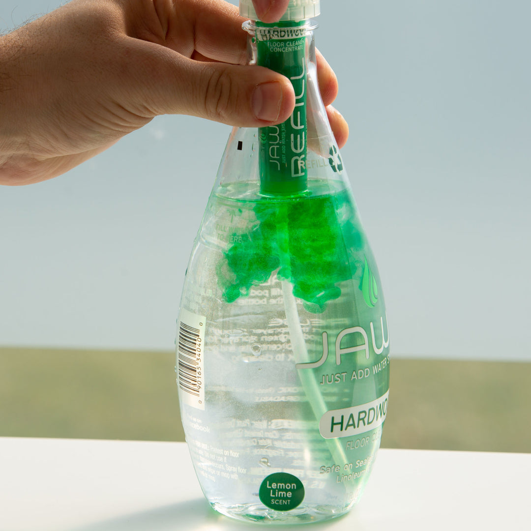 Hand holding a bottle of JAWS Hardwood Floor Cleaner with refill releasing green liquid into bottle against a neutral background.