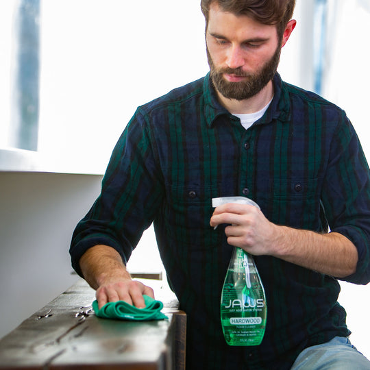 Man cleaning a wood surface with a green cloth and a bottle of JAWS Hardwood Floor cleaner.