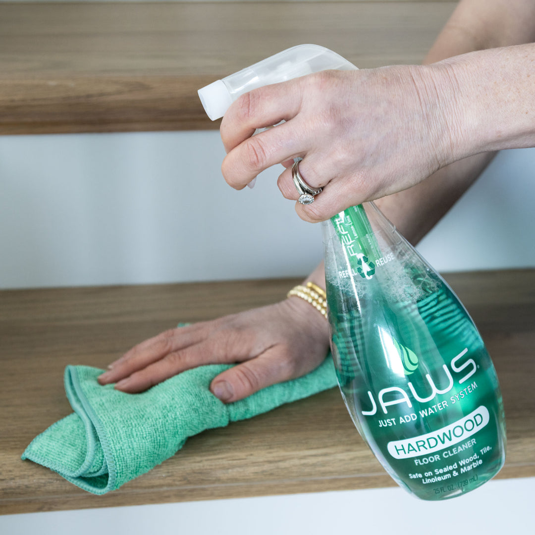 Person cleaning a wooden surface with JAWS Hardwood Floor Cleaner and a green microfiber cloth.
