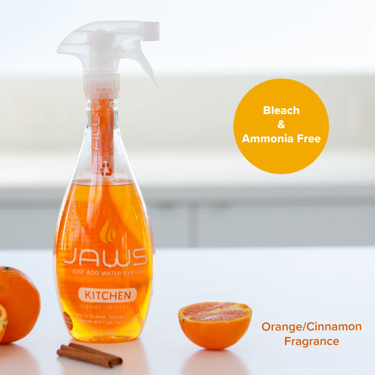 A bottle of JAWS Kitchen Cleaner with orange liquid, styled on a white surface with whole and sliced oranges, a cinnamon stick, and an orange circle badge that reads 'Bleach & Ammonia Free'.  Text overlay reads "Orange/Cinnamon Fragrance"