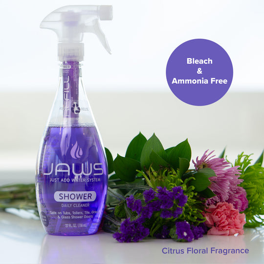 A bottle of Jaws Shower Daily Cleaner with a purple liquid and white spray top, shown next to a bouquet of purple and pink flowers. A purple circle reads 'Bleach & Ammonia Free' and text at the bottom says 'Citrus Floral Fragrance'.