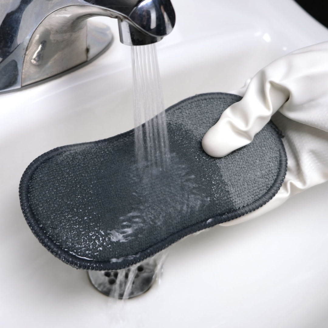 Gray cleaning pad being rinsed under running water in a sink with a white glove holding it.