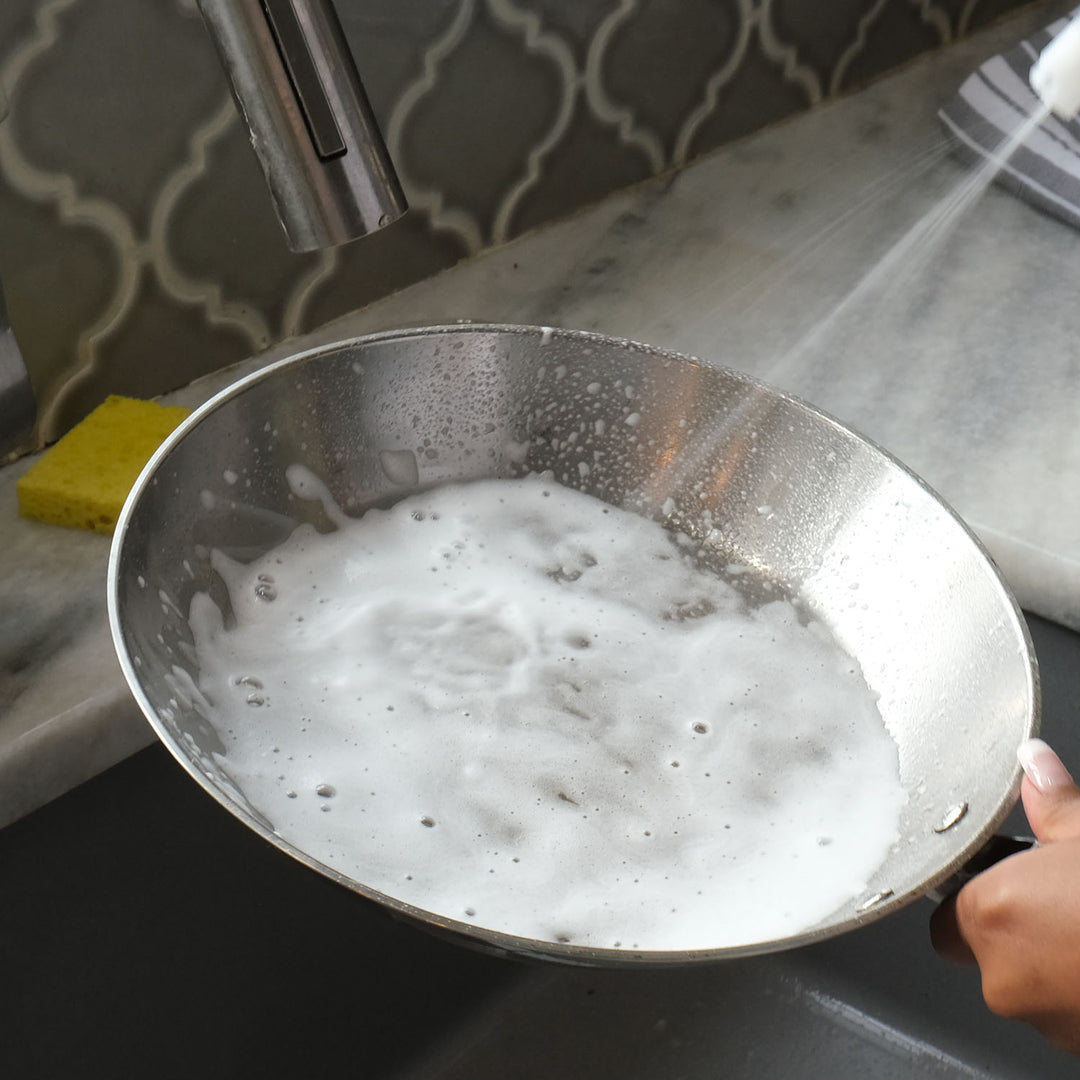 Stainless steel pan with soapy water being rinsed under a faucet.